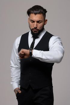 Bearded Hispanic guy in dark vest and white shirt with tie looking at camera with hands in pockets in studio