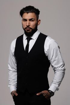 Bearded Hispanic guy in dark vest and white shirt with tie looking at camera with hands in pockets in studio