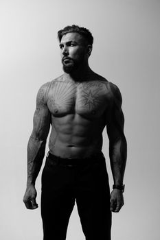 Hispanic shirtless male model with muscular tattooed torso standing with hands in pockets and looking away on gray backdrop