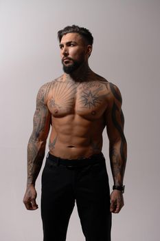 Hispanic shirtless male model with muscular tattooed torso standing with hands in pockets and looking away on gray backdrop