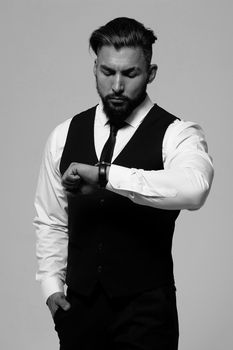 Bearded Hispanic guy in dark vest and white shirt with tie looking at camera with hands in pockets in studio