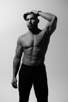 Hispanic shirtless male model with muscular tattooed torso standing with hands in pockets and looking away on gray backdrop