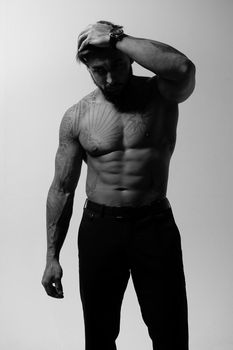 Hispanic shirtless male model with muscular tattooed torso standing with hands in pockets and looking away on gray backdrop