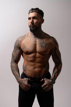 Hispanic shirtless male model with muscular tattooed torso standing with hands in pockets and looking away on gray backdrop