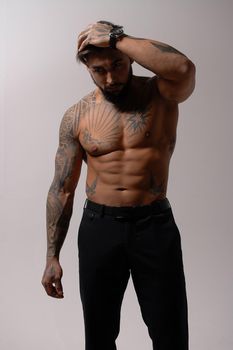 Hispanic shirtless male model with muscular tattooed torso standing with hands in pockets and looking away on gray backdrop