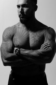 Hispanic shirtless male model with muscular tattooed torso standing with hands in pockets and looking away on gray backdrop