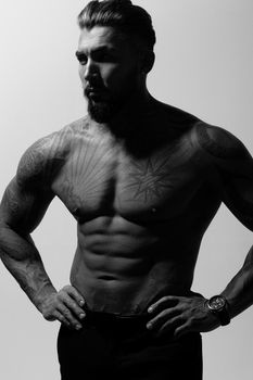 Hispanic shirtless male model with muscular tattooed torso standing with hands in pockets and looking away on gray backdrop