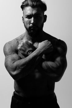 Hispanic shirtless male model with muscular tattooed torso standing with hands in pockets and looking away on gray backdrop