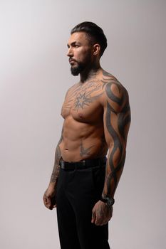 Hispanic shirtless male model with muscular tattooed torso standing with hands in pockets and looking away on gray backdrop