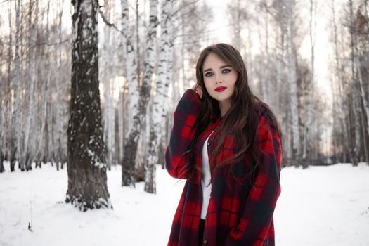 Charming female with red lips and in checkered shirt touching hair while standing in snowy winter woods and looking at camera