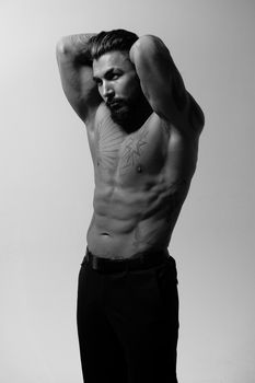 Hispanic shirtless male model with muscular tattooed torso standing with hands in pockets and looking away on gray backdrop