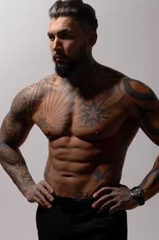 Hispanic shirtless male model with muscular tattooed torso standing with hands in pockets and looking away on gray backdrop