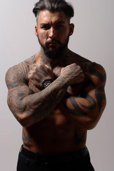 Hispanic shirtless male model with muscular tattooed torso standing with hands in pockets and looking away on gray backdrop