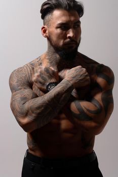 Hispanic shirtless male model with muscular tattooed torso standing with hands in pockets and looking away on gray backdrop