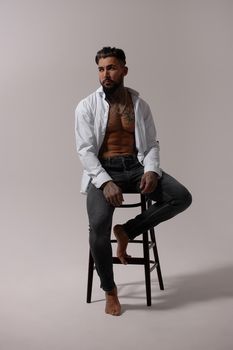 Hispanic shirtless male model with muscular tattooed torso standing with hands in pockets and looking away on gray backdrop