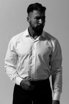 Bearded Hispanic guy in dark vest and white shirt with tie looking at camera with hands in pockets in studio