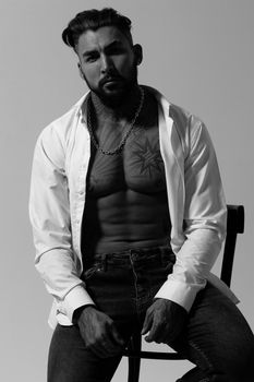 Hispanic shirtless male model with muscular tattooed torso standing with hands in pockets and looking away on gray backdrop