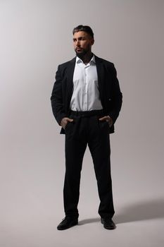 Bearded Hispanic guy in dark vest and white shirt with tie looking at camera with hands in pockets in studio