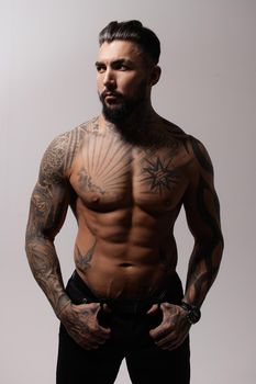 Hispanic shirtless male model with muscular tattooed torso standing with hands in pockets and looking away on gray backdrop