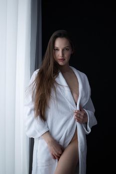 Low angle side view of sexy female with bare breast standing near window in room and looking at camera
