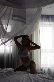Side view of seductive female in white lingerie sitting on soft bed and touching long ginger hair while looking at camera