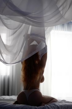 Back view of sexy female with long red hair taking off bra while sitting on soft bed at home
