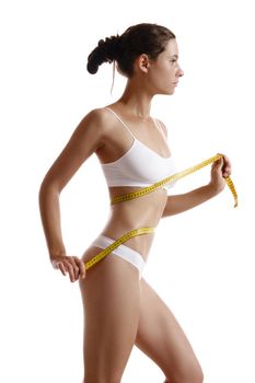 Perfect, slim, toned, young body of an alluring maiden in white underwear measuring herself with a measuring tape while posing sideways isolated on white. Beauty treatment. Plastic surgery and aesthetic cosmetology concept. Augmentation or reduce of a breast, cellulite removal. High resolution, close-up shot.