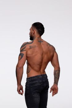 Hispanic shirtless male model with muscular tattooed torso standing with hands in pockets and looking away on gray backdrop
