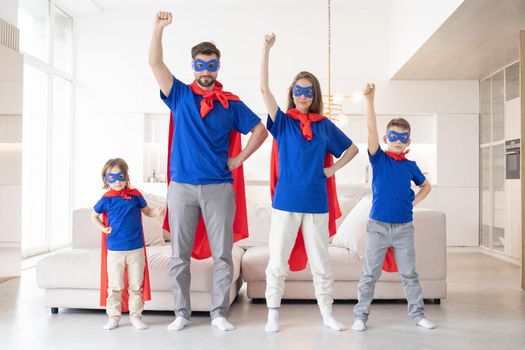 Full body of cheerful parents with two children in casual clothes and red superhero cloaks playing heroes at home