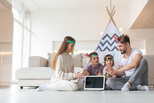 Happy family of parents and two children playing indian at home, wigwam tent, toy horse, feather roach