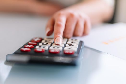 Close-up of a young woman's hand typing on her calculator as she works. Doing financial mathematical operations on a calculator. Young businesswoman and entrepreneur working in office or at home in white shirt.