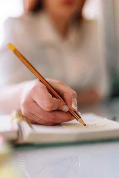 Close-up of the hand of a young girl in a white shirt writing with a pencil in her diary or notebook. Young entrepreneur and businesswoman teleworking. Young girl taking notes while studying. Woman studying.