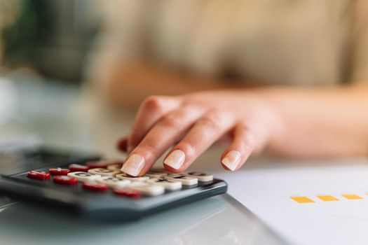 Close-up of a young woman's hand typing on her calculator as she works. Doing financial mathematical operations on a calculator. Young businesswoman and entrepreneur working in office or at home in white shirt.