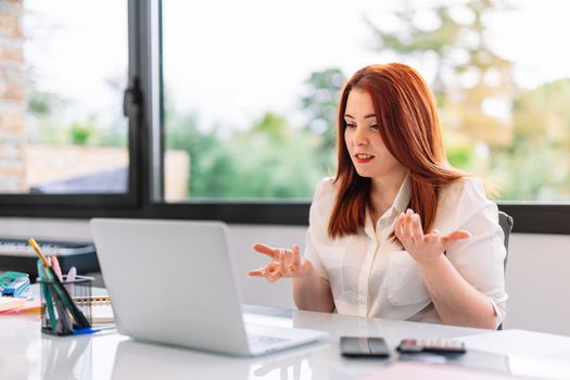 Pretty young redheaded pretty woman teleworking from home in white shirt. She is making a work video call explaining something. Very bright environment with a large window behind and natural light. Young businesswoman and entrepreneur working on white laptop.