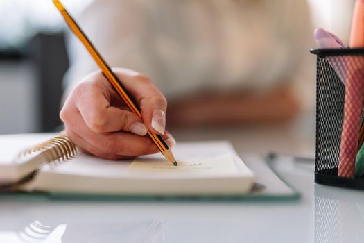 Close-up of the hand of a young girl in a white shirt writing with a pencil in her diary or notebook. Young entrepreneur and businesswoman teleworking. Young girl taking notes while studying. Woman studying.