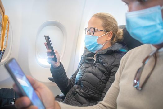 A young couple wearing face mask, using smart phone while traveling on airplane. New normal travel after covid-19 pandemic concept