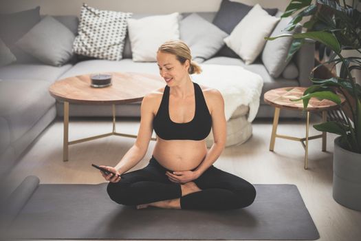 Pregnancy and technoloy supported healthy lifestyle concept. Cheerful happy pregnant woman using smart phone application while exercising on yoga mat on living room floor at home