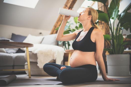 Young happy and cheerful beautiful pregnant woman taking a break, hydrating, drinking water from the botle after home well being workout progrem