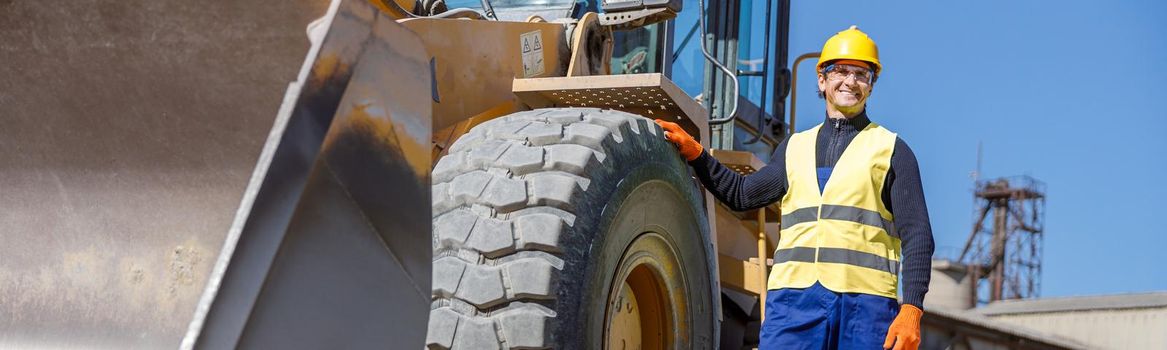Joyful matured man factory worker placing hand on wheel and smiling while standing near tractor