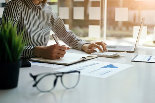 businesswoman or accountant taking note and working on calculator and laptop computer to calculate business data during make note at notepad, accountancy document at office.