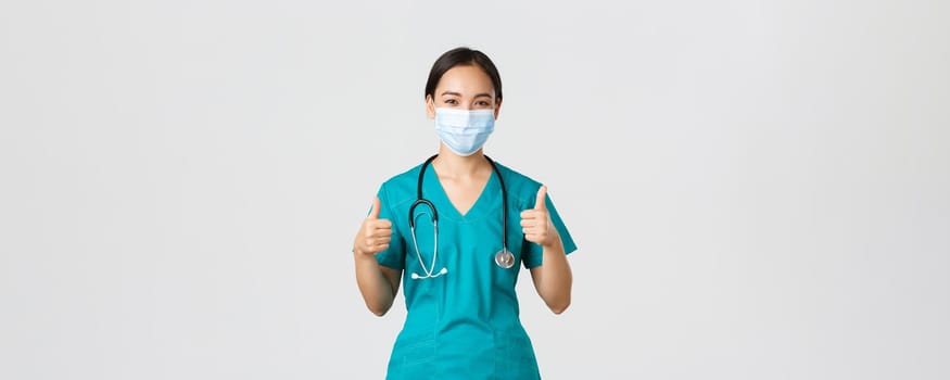 Covid-19, coronavirus disease, healthcare workers concept. Confident, smiling asian doctor, female nurse in scrubs and medical mask profive good quality service, show thumbs-up in approval.