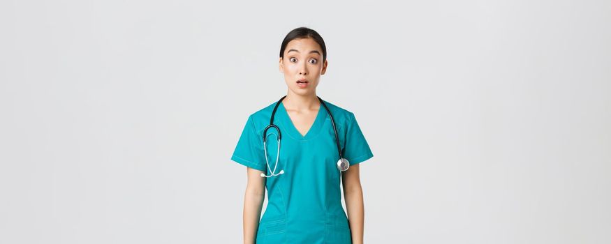 Covid-19, healthcare workers, pandemic concept. Impressed and surprised asian nurse in scrubs drop jaw and stare at camera speechless, looking amazed over white background.