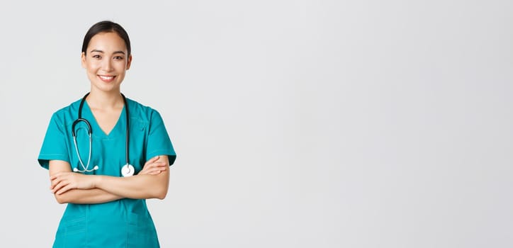 Covid-19, healthcare workers, pandemic concept. Portrait of confident smiling, attractive asian female nurse in scrubs, with stethoscope, cross arms chest and looking at camera, white background.