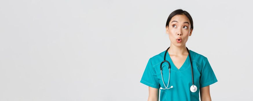 Healthcare workers, preventing virus, quarantine campaign concept. Impressed and amazed asian female doctor, nurse in medical scrubs looking upper left corner pleased, white background.