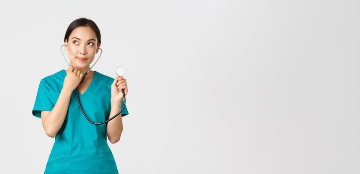 Covid-19, healthcare workers and preventing virus concept. Cute smiling asian female intern learn how listen lungs with stethscope, doctor examine patient, looking thoughtful upper left corner.