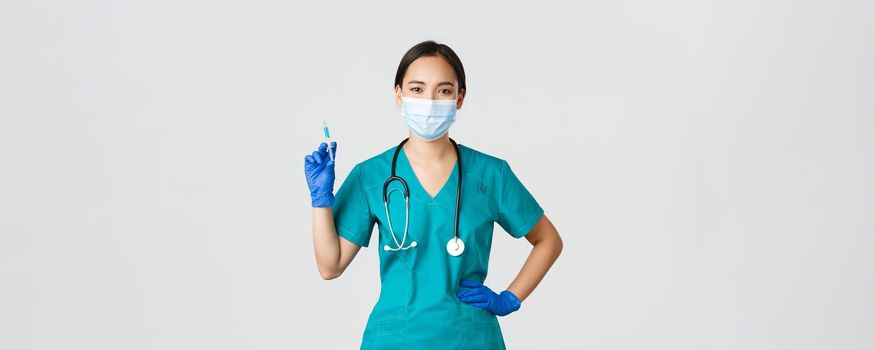 Covid-19, coronavirus disease, healthcare workers concept. Confident asian female nurse, doctor in medical mask and rubber gloves, holding syringe with vaccine, standing white background.