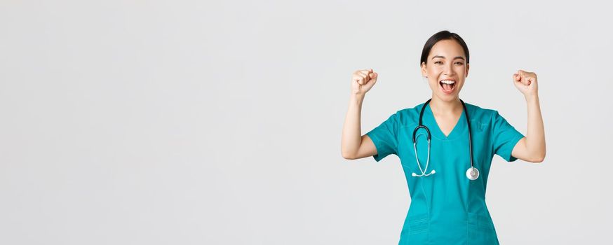 Covid-19, healthcare workers and preventing virus concept. Successful confident asian female doctor or nurse in scrubs fist pump and shouting yes, rejoicing, encourage herself, aim for victory.