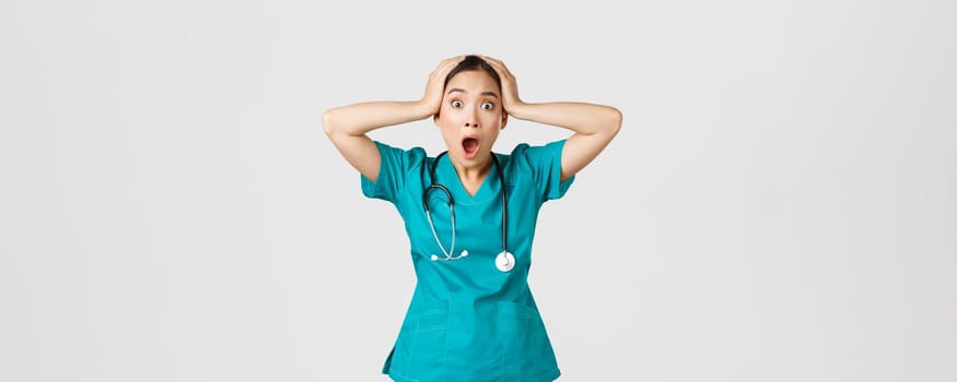 Covid-19, healthcare workers and preventing virus concept. Shocked and anxious asian female doctor in panic, grab head and screaming alarmed, have big problem, got in trouble, white background.