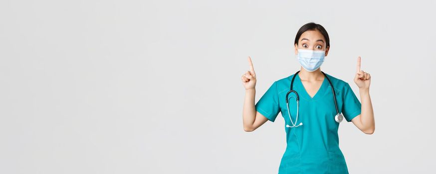 Covid-19, coronavirus disease, healthcare workers concept. Intrigued asian female doctor, physician or intern in medical mask and scrubs, looking curious, pointing fingers up, white background.
