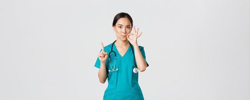 Covid-19, healthcare workers, pandemic concept. Serious-looking worried asian female nurse, physician asking keep secret, shaking finger and showing mouth seal, zipping lips gesture.